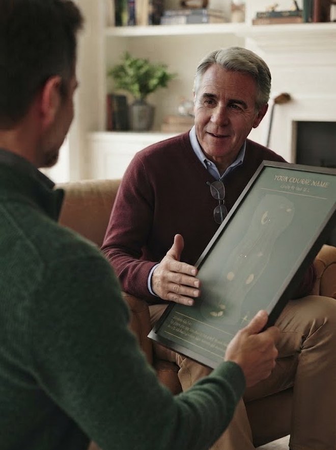 A son handing his father a framed Tee Sheetz course-map poster in a living room